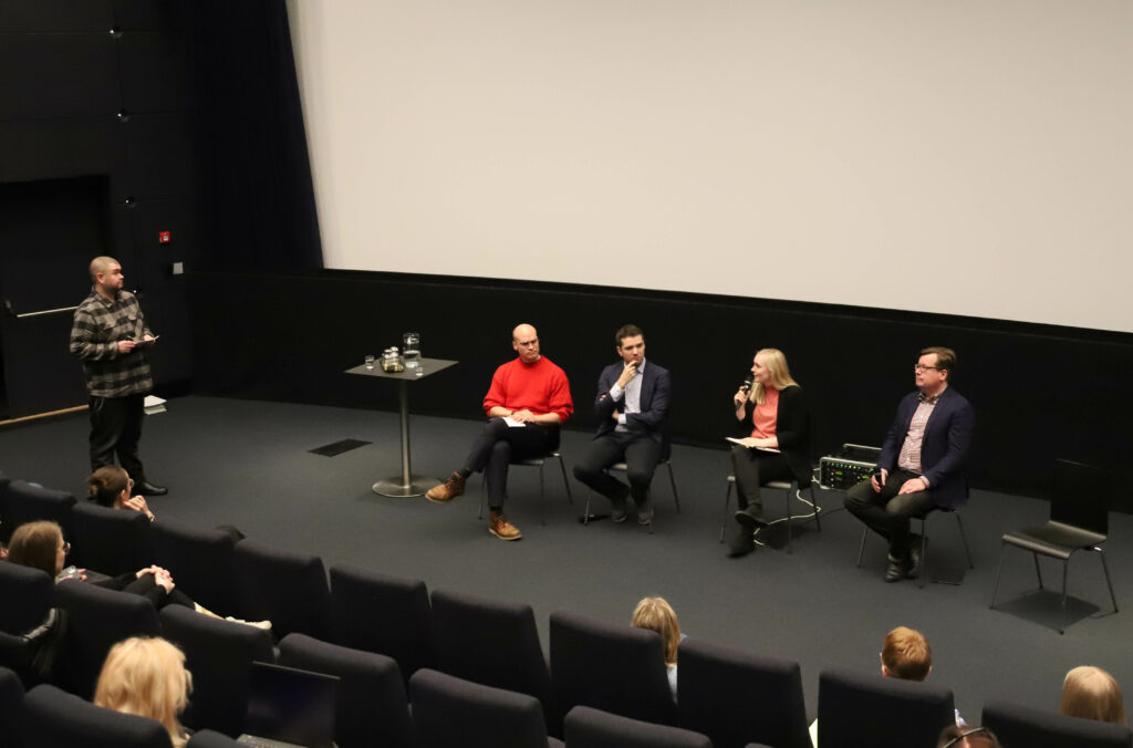 Kuvassa paneelikeskustelu lavalla katsomon edessä. Paneelia juontaa Juha Kahila, jonka oikealla puolella istuvat panelistit Riku Nieminen, Ted Apter, Maria Ohisalo ja Mikko Aaltonen.